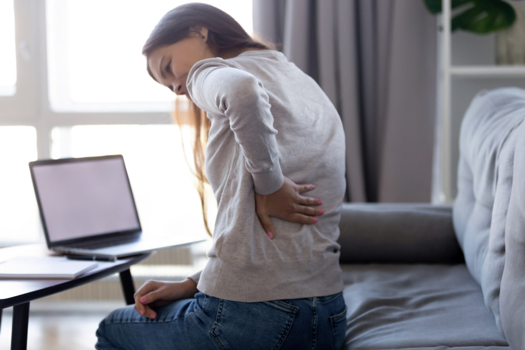 woman facing her laptop sitting on a couch feel pain in her lower back